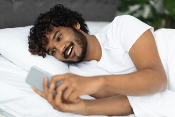 Smiling hindu guy laying in bed with smartphone