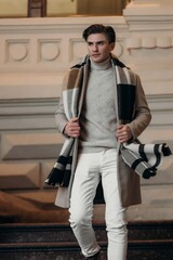  Young man wearing coat walking in the street. Young bearded guy with modern hairstyle in urban background