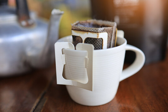 Sachet Or Hanging Drip Coffee The Ear Was Placed In A White Cup On A Wooden Floor, With A Kettle Laid In A Blurry Background.