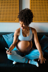Happy fit pregnant woman at home texting on smartphone. Afro hairstyle black female using her phone. - 489013412