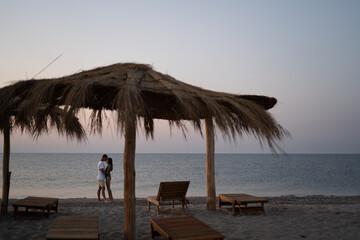 Young happy loving couple on the beach under straw umbrellas hugging at sunrise or sunset. Honeymoon.