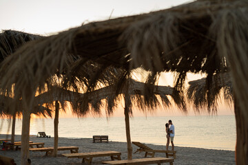 Young happy loving couple on the beach under straw umbrellas hugging and kissing at sunrise or sunset. Honeymoon.