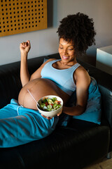 Relaxed young black pregnant woman at home eating an organic healthy salad bowl in her bed.