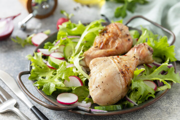 Healthy clean eating. Meat with fresh salad. Baked chicken drumsticks and a vegan salad made from a of green leaves and spring vegetables on a stone countertop.