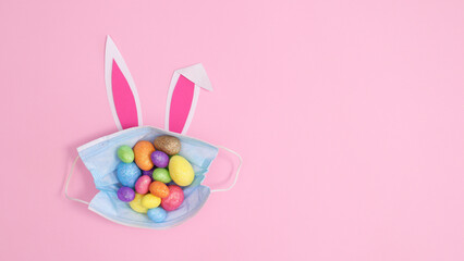 Face mask with colorful Easter eggs and bunny ears on pastel pink background. Creative copy space flat lay