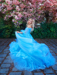 Fantasy happy woman princess in long fluffy blue dress, like Cinderella, circles in dance, fabric flies waves in motion. Fairy girl goddess blond hair. Spring blooming nature, sakura tree pink flowers © kharchenkoirina