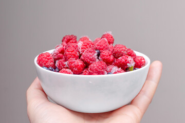 Close-up of frozen raspberries in a bowl held by a man's hand on a neutral gray background. Delicious berry dessert