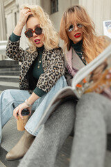 Fototapeta na wymiar Fashionable beautiful women in stylish retro clothes sit in the city on the steps, drinking coffee and reading a magazine