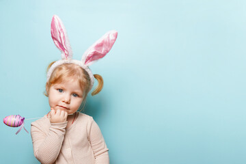 Obraz na płótnie Canvas A little girl with bunny ears on her head holds easter egg on blue background. Easter and spring symbol