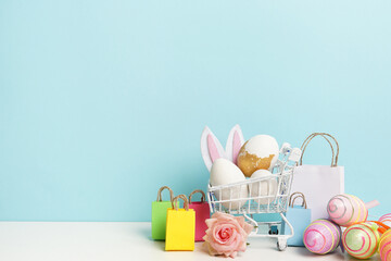 Easter shopping cart with colorful paper bags, bunny ears and eggs on blue background. Copy space