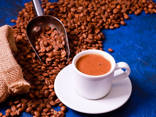 Espresso white mug, coffee beans scattered on blue concrete background table from rustic sack, metal scoop. Coffee shop, food, roaster, morning drink concept. Close up