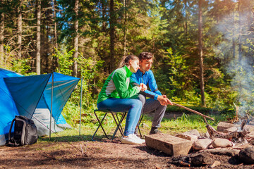 Naklejka na ściany i meble Young couple of travelers enjoy campfire in summer forest. Tourists sitting by tent hugging. Camping in woods