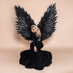 Elegant fashion. Stunning blonde woman in elegant long black dress and big wings on beige...