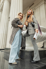 Two fashion pretty young woman with coffee cup and magazine in stylish autumn clothes with leopard jacket, jeans, elegant suit and shoes stands and poses in the city