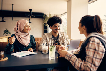 Happy Muslim student uses laptop while studying with female friends at university cafeteria.