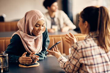 Young Islamic woman and her female friend looking at something on cell phone in a cafe.