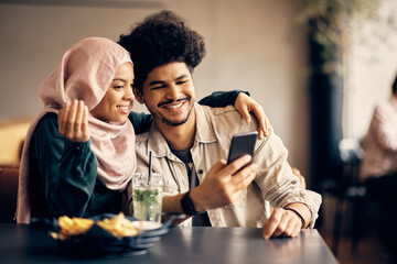 Happy Muslim man and his girlfriend use smart phone in cafe.