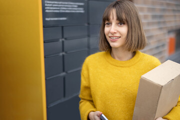 Happy woman with parcel and phone standing near automatic post terminal outdoors. Concept of contactless and smart delivery. Idea of modern shipping and logistics. Woman wearing yellow sweater