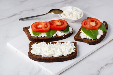 Slice of rye bread with cottage cheese and tomatoes on a wooden cutting board on a white background