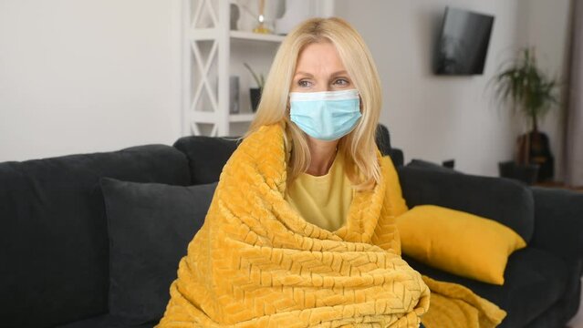 Senior sick woman wearing protective face mask feeling unwell at home, sits on the couch covered with blanket, feels chills and fever, a female has disease and self isolated at home
