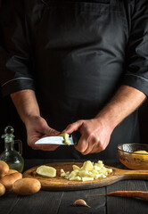 The chef cuts raw potatoes into pieces with a knife before preparing breakfast or dinner. Close-up of a cook hands while working in a restaurant kitchen.