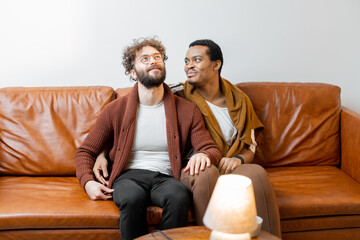 Gay couple with different nationality having close conversation while sitting on a couch at home. Concept of homosexual relations and lifestyle at home. Caucasian and hispanic man together indoors