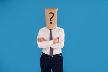 Businessman with brown paper bag on head with question mark, standing on blue studio background