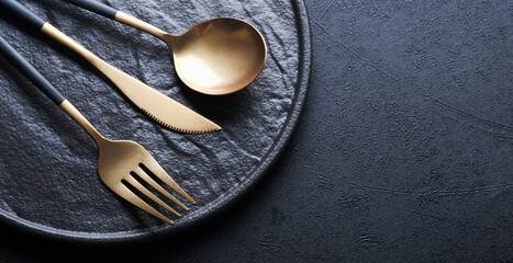 Gold spoon, fork, knife and clean empty black plate. Set of stylish black and gold cutlery on black background. Fashionable and luxury eating. Top view. Copy space for your text.