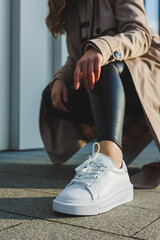 Stylish trendy white women's leather sneakers on the pavement on a sunny day. Women's legs in...