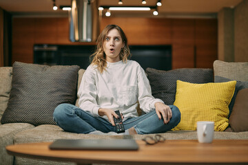 Surprised young woman watching serials or movie, sitting on sofa, saying wow, wondered by...
