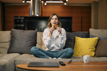 Surprised young woman watching interesting movie or series, eating fastfood, sitting on couch at home