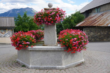 Fototapeta premium stone water fountain with colorful flower pots in the plaza, alps, france