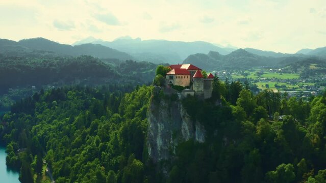 Bled castle built on high cliff against giant mountains in Slovenia. Bled lake with blue water surrounded by lush green trees in summer. Panorama view