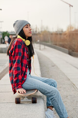 Young Hispanic woman sitting with skateboard on her knees. Venezuelan Latin woman with headphones.