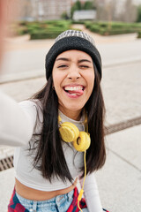 latina taking a selfie while making a funny face sticking out her tongue. Young Hispanic girl wearing casual clothes and a hat with headphones smiling. - obrazy, fototapety, plakaty