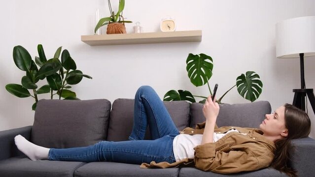 Calm young woman 20s lying relaxing on comfortable couch at home using modern smartphone, relaxed brunette girl rest on cozy grey sofa texting or chatting on cell, shopping online on cellphone