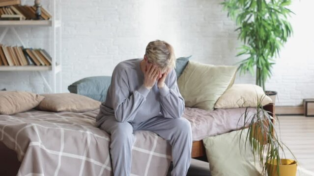 Male depression. An unhappy young man is sitting on the bed and thinking about problems, covering his face with his hands.