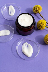 Petri dishes with white cream smears and cream in dark glass jar on violet background with yellow flowers of craspedia.