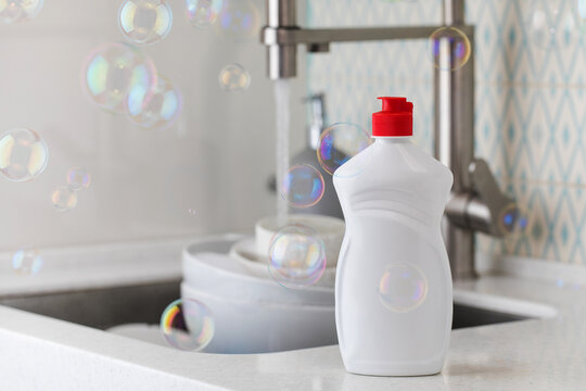 White bottle with dishwashing gel with soap bubbles around on the background of a sink with dirty dishes.