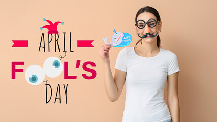 Young woman in funny disguise holding paper with text APRIL FOOLS on beige background