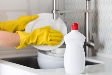 A white bottle with dishwashing gel on the background of a housewife washes dishes in a sink in...