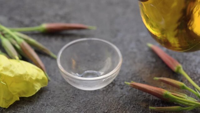 Pouring evening primrose oil from a bottle into a bowl, with fresh Oenothera biennis flowers, slow motion