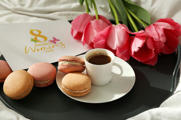 Tray with cup of coffee, sweet macaroons, greeting card and flowers for International Women's Day celebration on bed, closeup