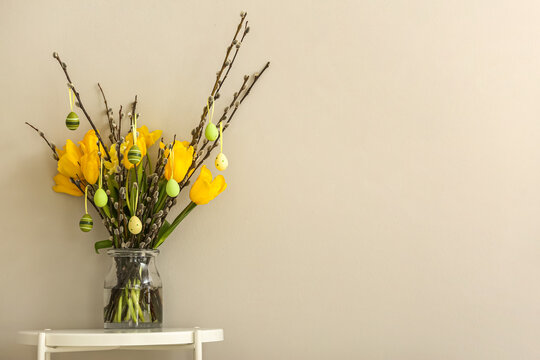 Vase with pussy willow branches, tulip flowers and painted Easter eggs on table near beige wall