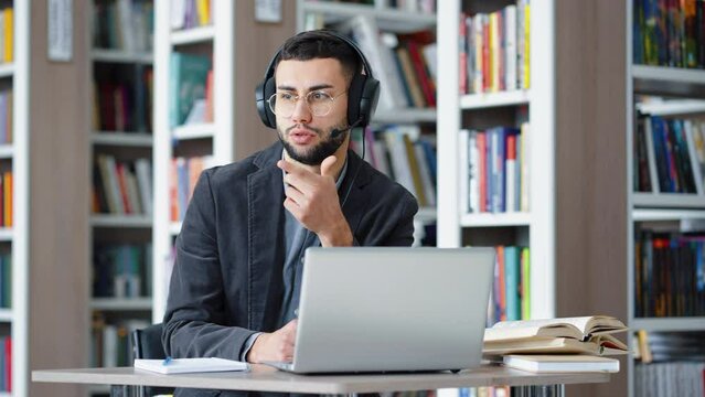 Bearded man with eyeglasses wearing headset talking and touching his chin, laptop standing on table in front of him, bookcases on blurred background. Distance learning, zoom event in library