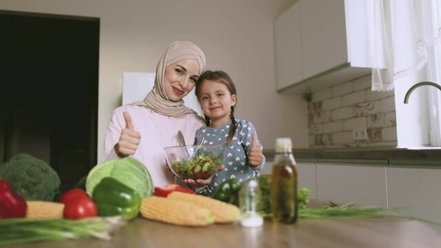 Head shot portrait smiling arabian muslim woman with caucasian daughter cooking salad together, sitting at wooden table in kitchen. Happy family preparing meal, enjoying weekend