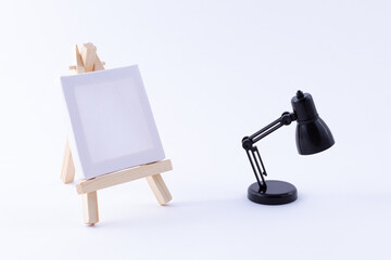 Wooden Easel Miniature with Blank White Square Canvas for Artists and Painters - Mockup. Mini Wooden Stand with Clean Artboard and Small Black Table Lamp on White Background, Copy Space