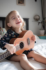 toddler girl playing ukulele guitar sitting on the bed at home