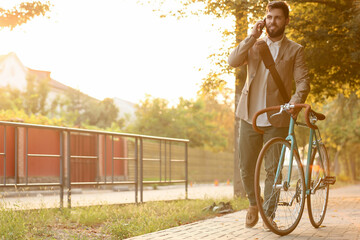Young bearded businessman with bicycle talking by mobile phone on city street