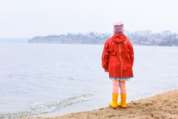Little girl in stylish rubber boots and raincoat walking near river